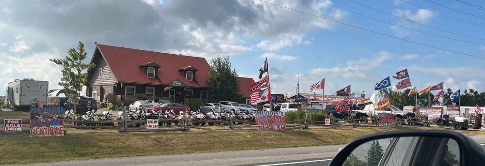Exterior photo of Treasure Island, taken from a car. There are at least four confederate flags flying, two half-Canadian, half-US flags, two Metis flags, and four KFN flags. And... India, I think? And many, many hand-painted signs, including on the roof of Willy's Man Cave.
