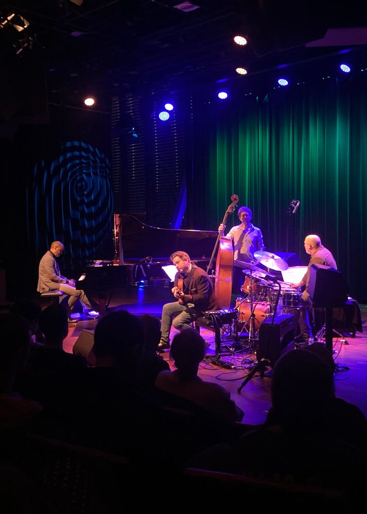 A jazz quartet on stage; piano, bass and drums take a breather, whilst the guitarist throws shapes.
