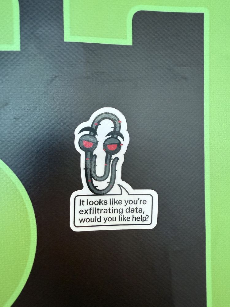 A sticker shaped like an evil-looking paperclip with red eyes, placed on a large green letter “R” on a black background. The sticker’s speech bubble says, “It looks like you’re exfiltrating data, would you like help?”
