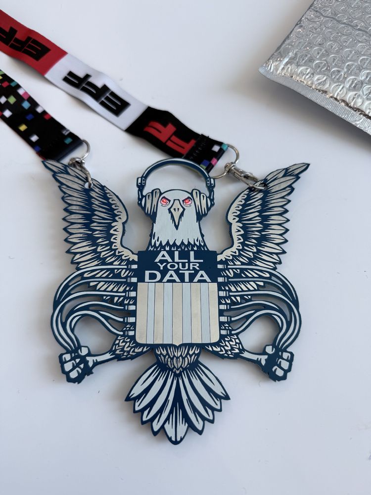 A conference badge shaped like an eagle with outstretched wings, wearing headphones, and red eyes. The eagle’s chest features a shield with the words “ALL YOUR DATA” above vertical stripes. The badge is attached to a lanyard with the EFF logo and colorful pixel patterns