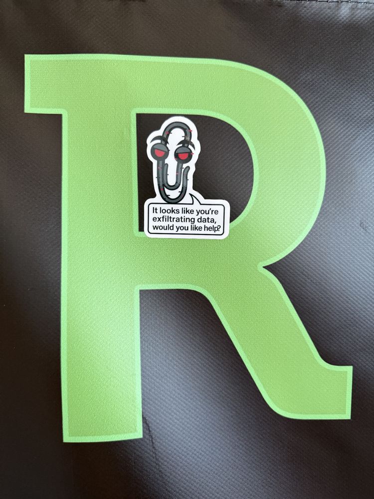 A sticker of a sinister-looking paperclip mascot with red eyes is placed on a large green letter “R” printed on a dark textured background. The sticker includes a speech bubble that says, “It looks like you’re exfiltrating data, would you like help?”