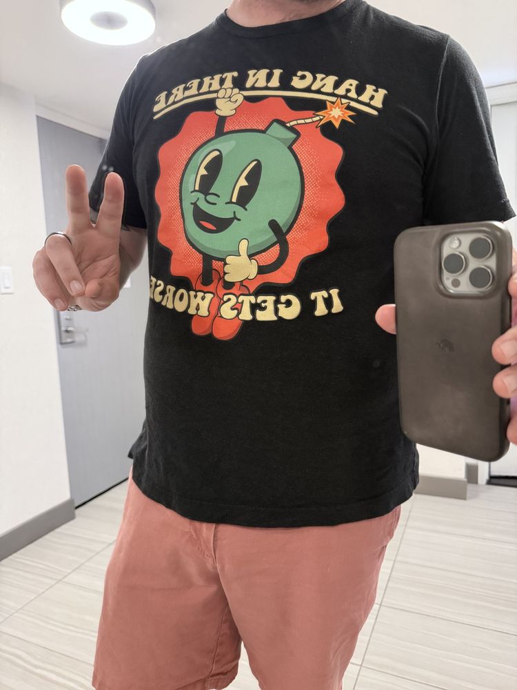Person taking a mirror selfie wearing a black T-shirt with a cartoon-style green bomb character smiling and giving a thumbs-up. The shirt text reads “HANG IN THERE” above the bomb and “IT GETS WORSE” below. The person is making a peace sign with their left hand and wearing salmon-colored shorts.