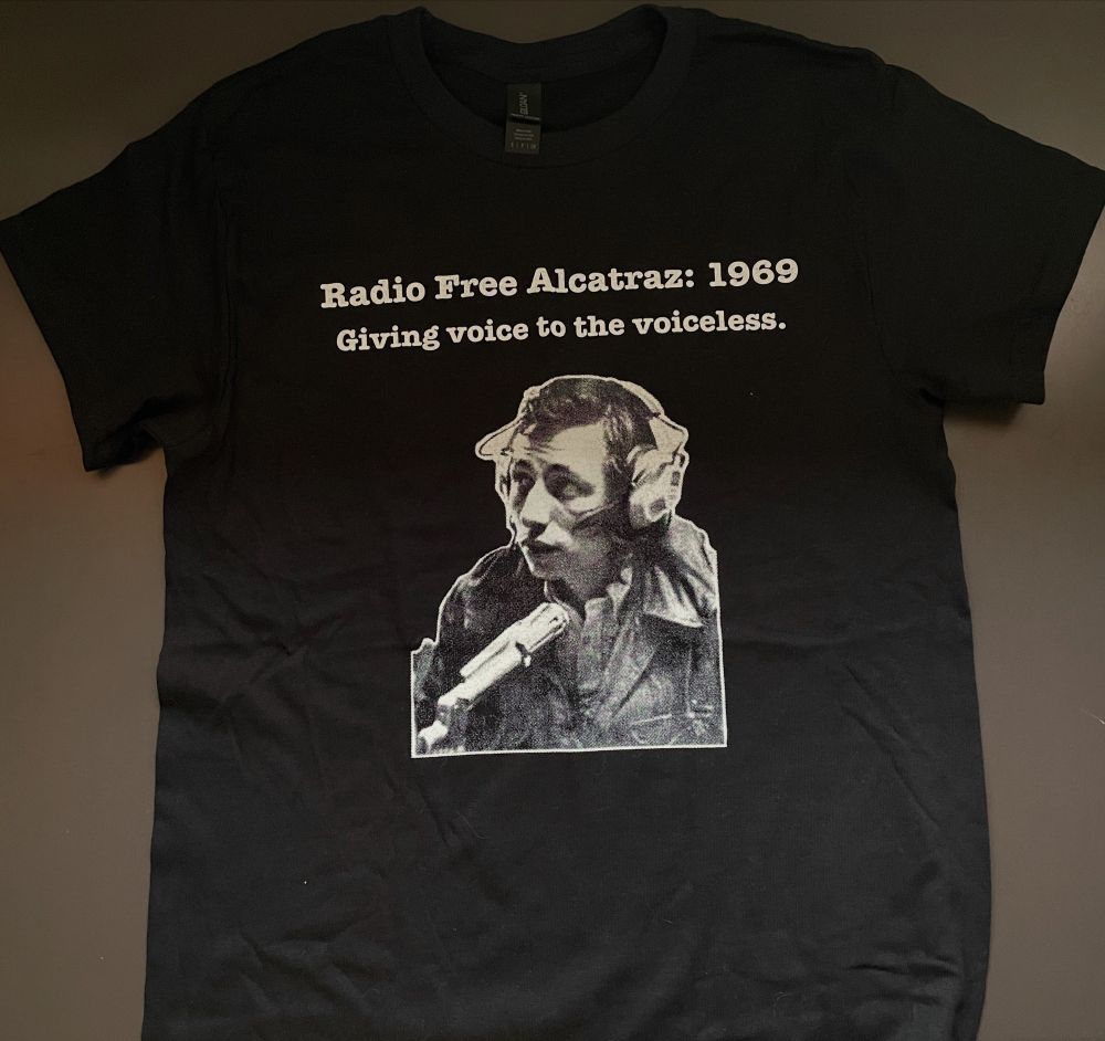 Black t shirt laying on a silver work table with white text that reads “Radio Free Alcatraz 1969 / Giving voice to the voiceless.”