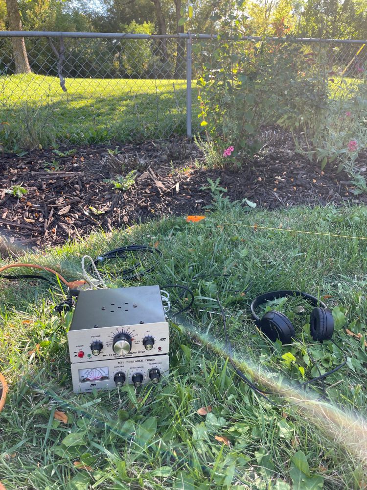 20 meter band silve radio stacked on top of a SWR meter sits on green grass in front of a flower bed. a long yellow wire antenna stretches across the yard beside it. a pair of black headphones are also lying on the ground.