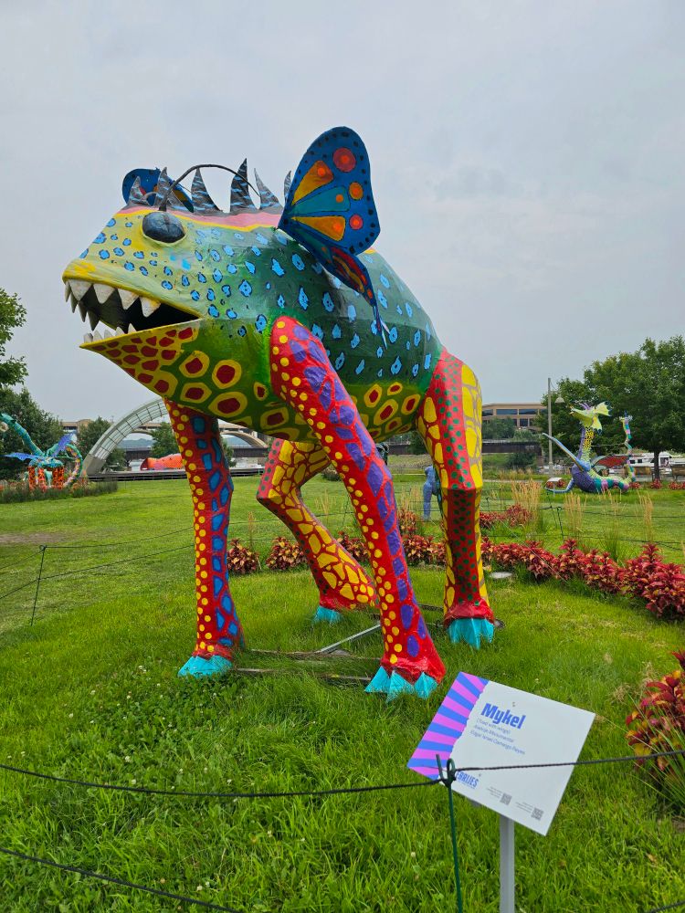 mykel: a statue of a colorful frog with green and blue on its body, red legs, pointy white teeth, and blue and orange butterfly wings.