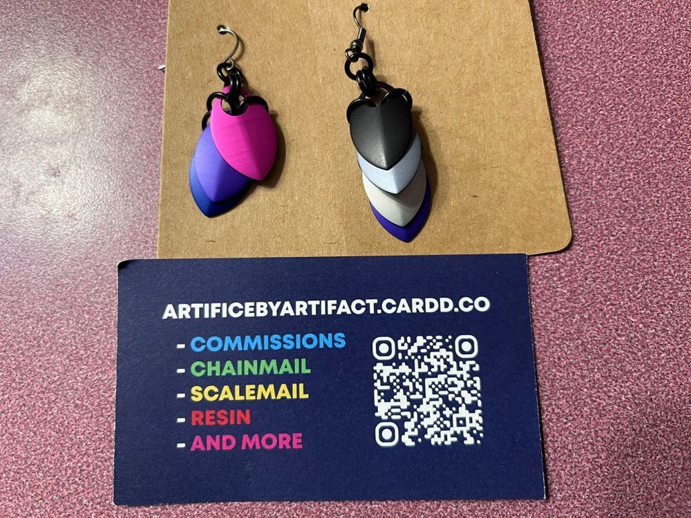 Image shows two earrings made of dangling scalemail: the one on the left is the bi flag, the one on the right is the ace flag. A business card below is for artificebyartifact.cardd.co.