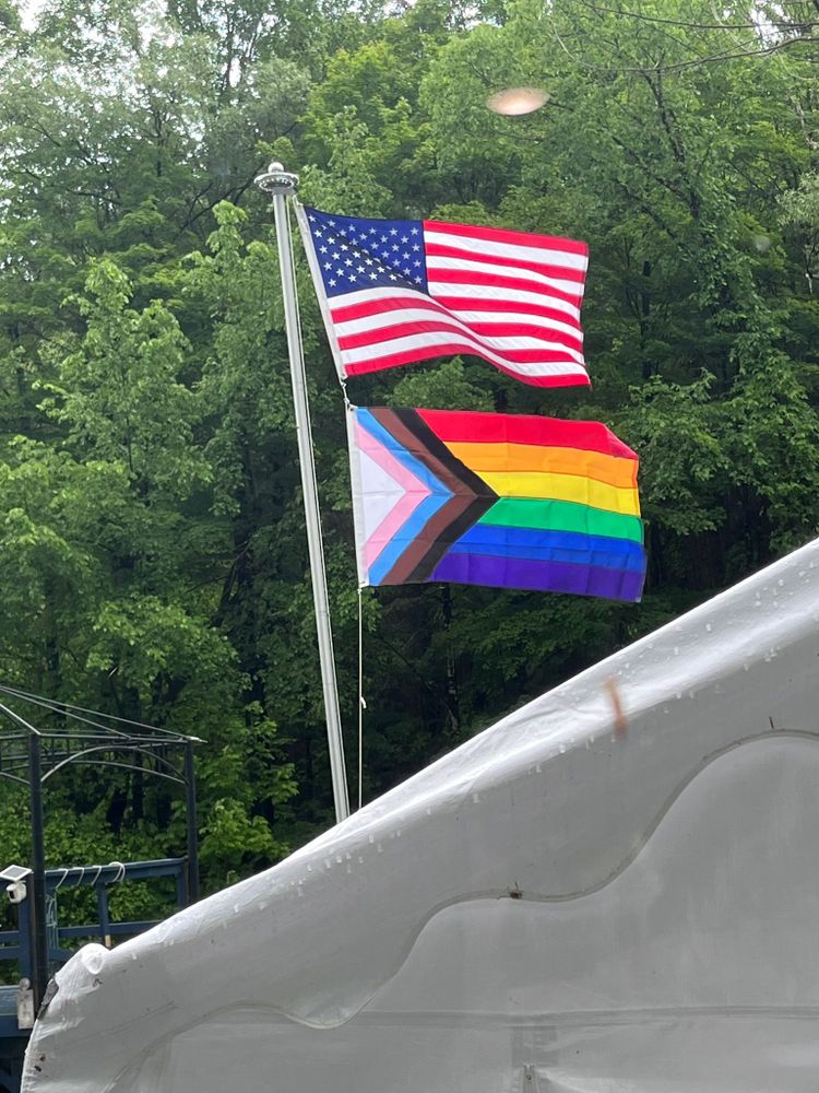 A flag pole with two flags flying in the rain. The one on top is the US flag, and the one below is the progress pride flag.