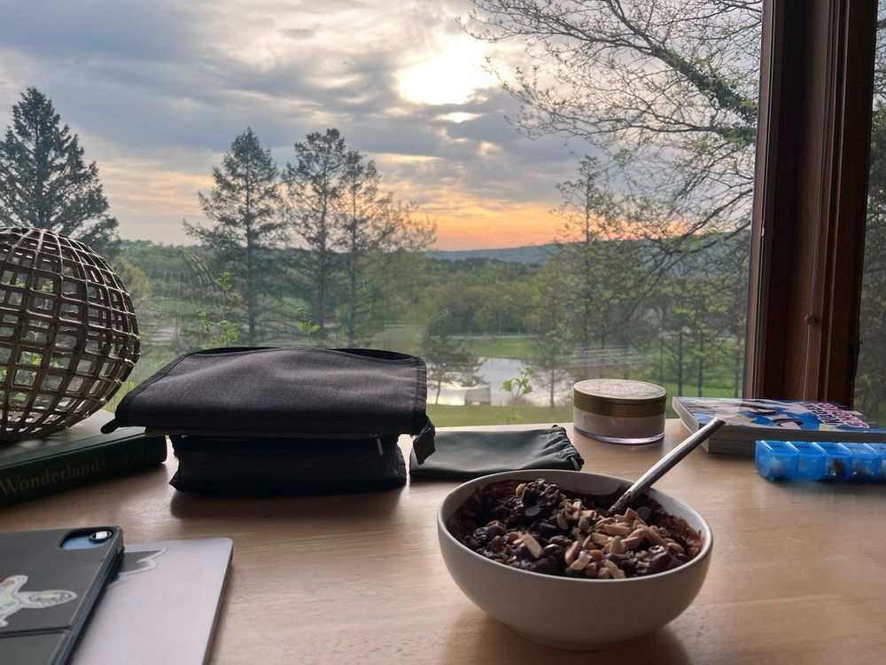 A sunrise in streaked orange and clouds through a window past a desk cluttered with stuff and a bowl of oatmeal. 
