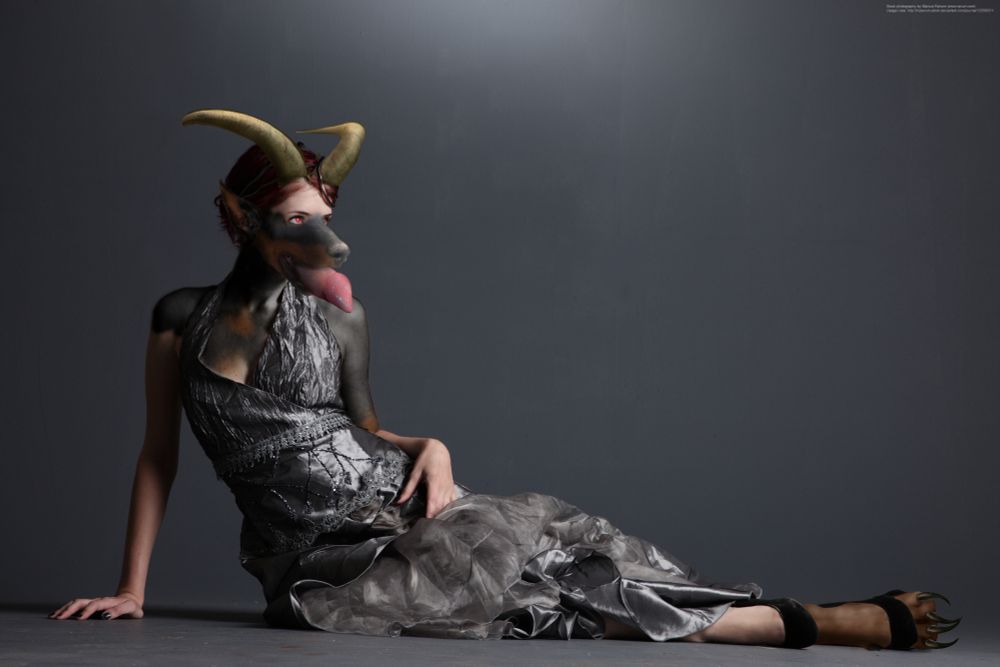 A photomanipulation of a woman in a gray dress, lying on the floor as she transforms into an anthro Doberman demon, with curved horns, red eyes and a large canine cocktongue