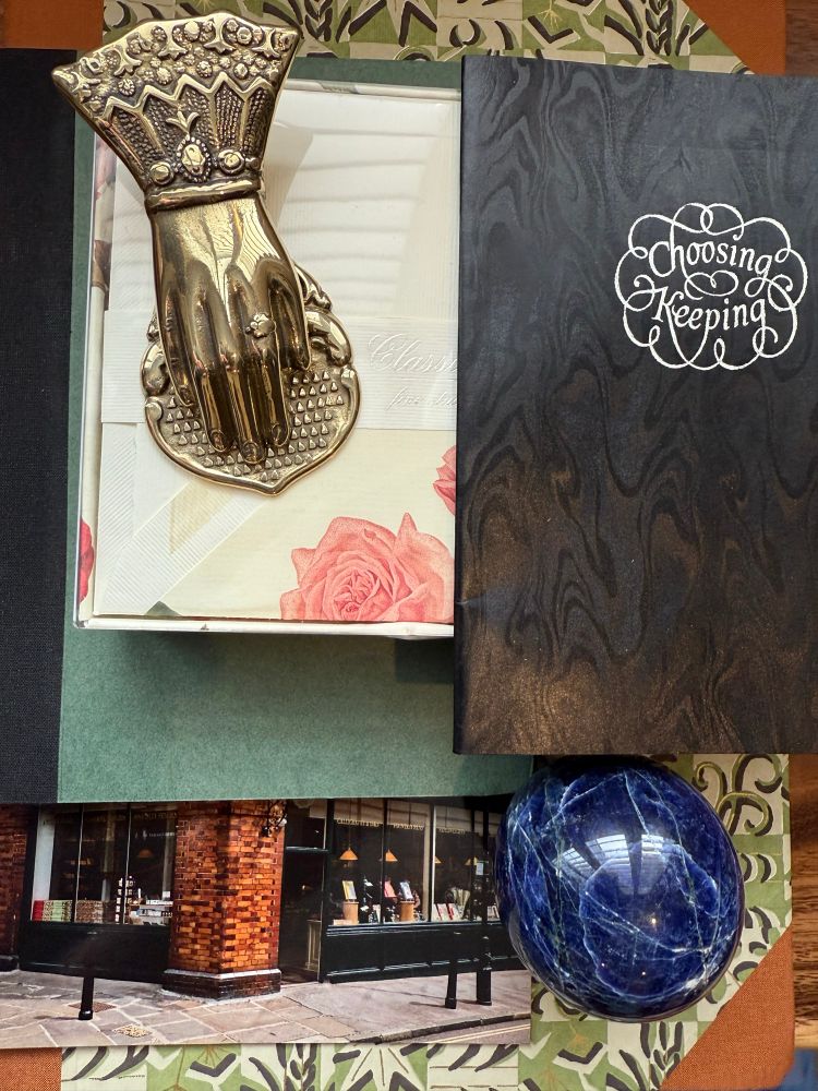 Items from the stationery store called Choosing Keeping. A “hand” clip sits on top of a box of rose stationery, along with a small black journal. This is all on top of a green notebook and their post card. All of this is on top of their giant A4 size patterned paper covered journal. There is a sodalite sphere on the bottom right hand corner. 