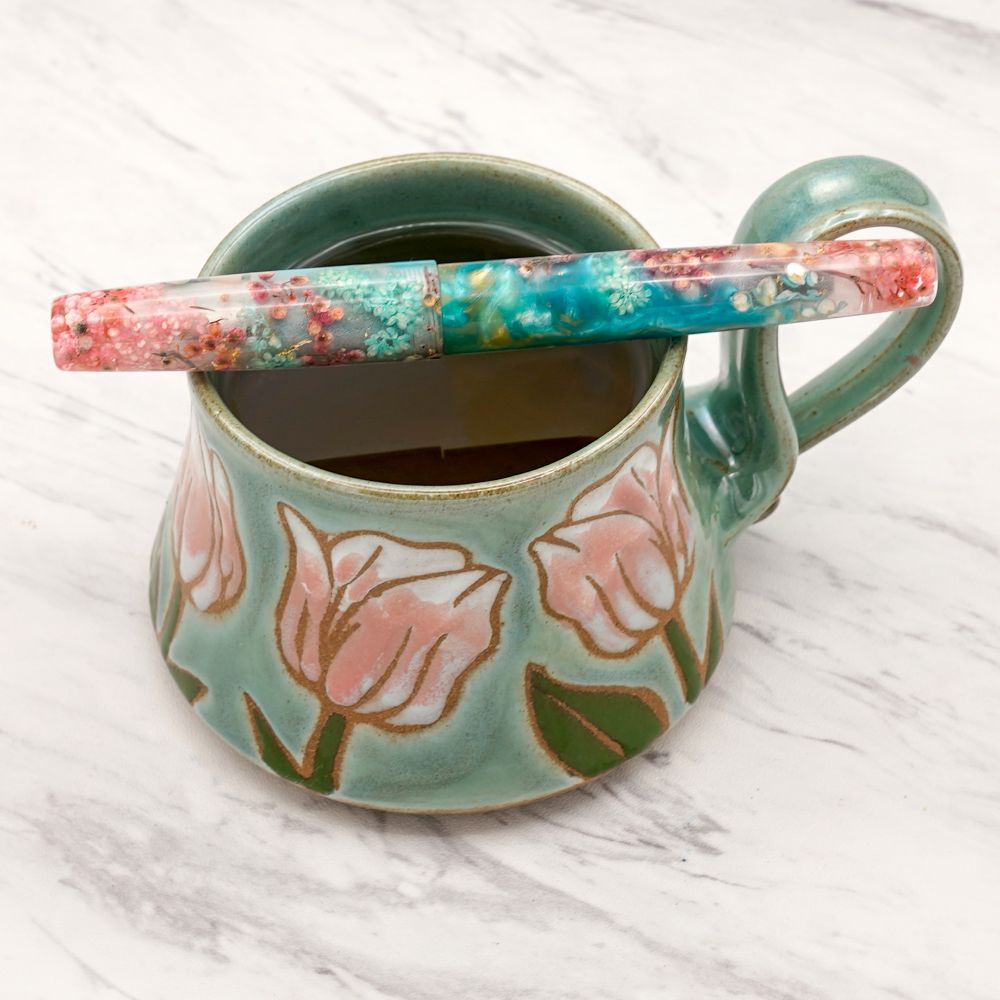 Fountain pen with turquoise swirls and embedded pink flowers on top of a turquoise and pink tulip motif mug