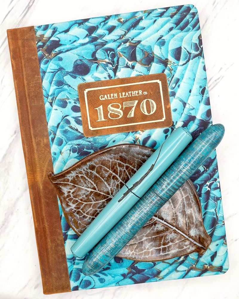Two blue urushi pens in different styles on a ceramic leaf dish on top of a marbled paper notebook in shades of turquoise with leather binding 