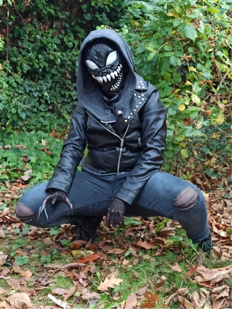 A photo of the artist in a crude cosplay of her persona, Voidling. She's a small statured person, crouching on the ground surrounded by greenery and dry leaves. She's dressed all black, with leather boots, ripped jeans, a faux leather jaket and a hoodie with the large hood thrown on. Visible skin is painted black and the face is concealed under a bootleg venom halloween mask, that gives a close enough aproximation to Voidling's face. Left hand has long black nails glued on to simulate claws. The person is looking at the camera, the mask giving a wide toothy grin. 
