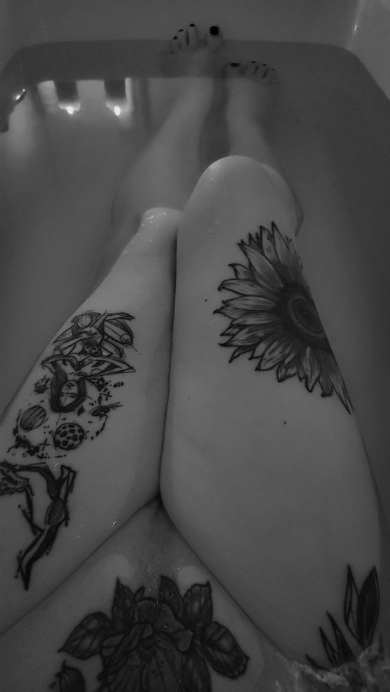 A monochrome image of the legs and feet of a transgender woman showing several large tattoos on her legs and mons. She has almost completely shaved off her pubic hair, with only a small strip remaining 