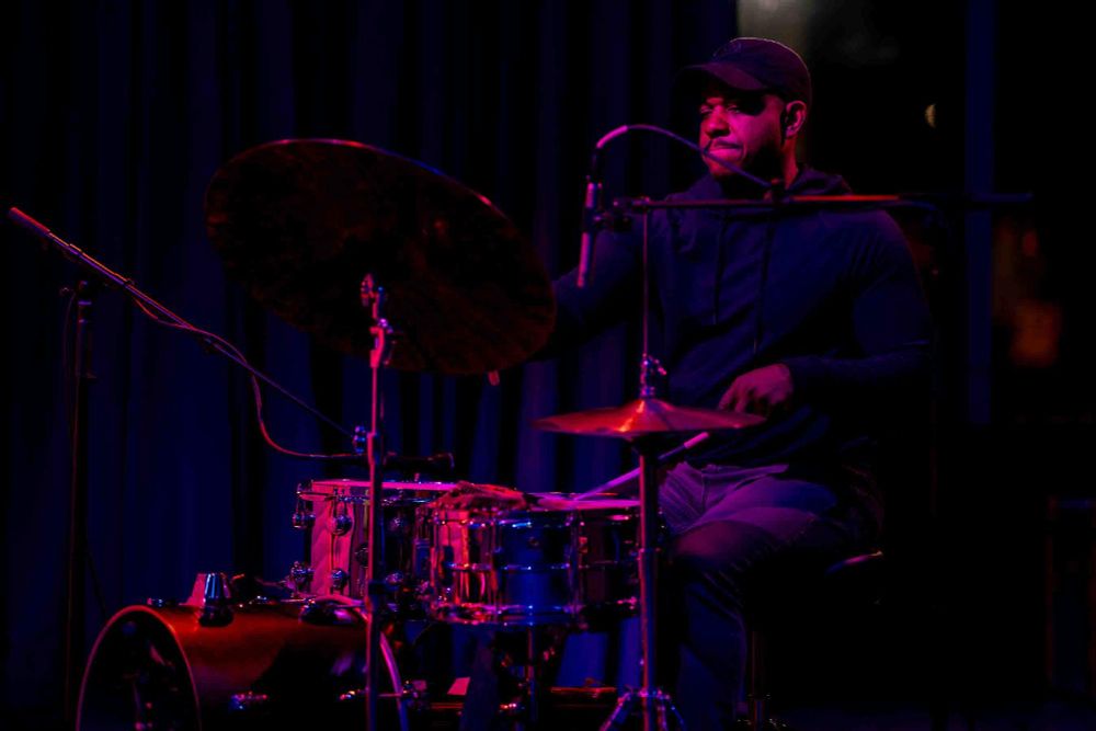 A musician is performing on a drum set, illuminated by pink stage lighting.
