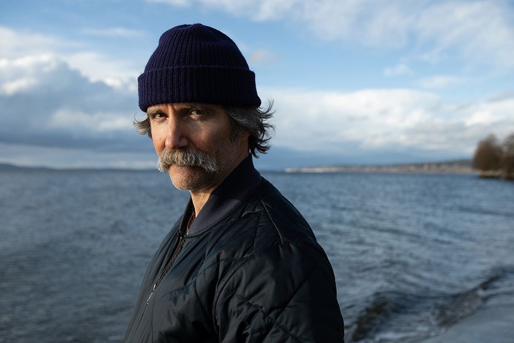 Person in a purple beanie and blue jacket standing by a lake with clouds in the sky.
