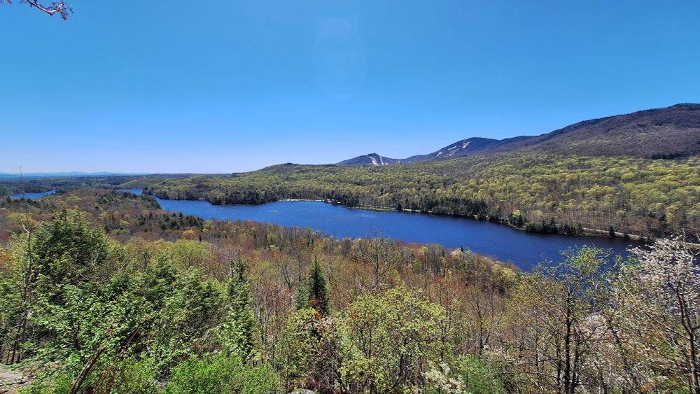 A wide-angle view of a valley in Mount Orford National Park (Quebec, Canada) with a blue lake in the centre and a hilly ridge in the background. The bright green leaves on the trees all around the lake have just started to grow (photo taken early May). There is still some snow from the winter on the ski slopes in the background.