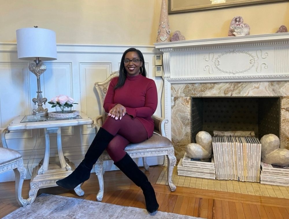 A woman wearing a dress and tights in a dark shade of red and black boots is seated while posing for a picture.