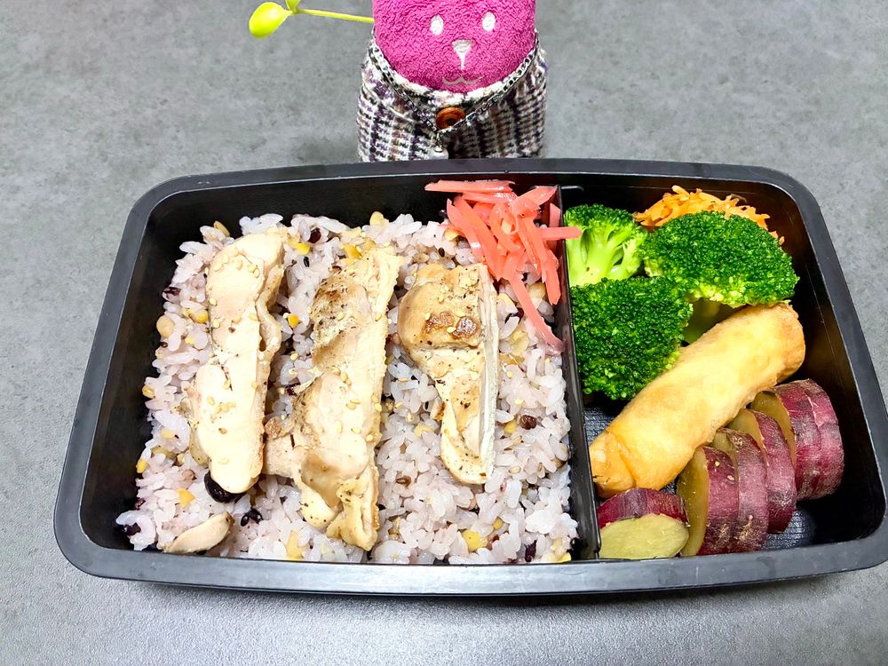 雑穀米の上にベニショ、鶏もも肉、おかずにブロッコリー、春巻、にんじんナムル、さつまいもの入った弁当、隣にばにちゃ