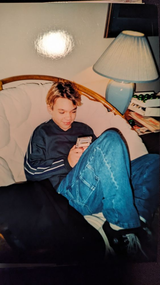 Me, only just high school age, curled up in a papasan, playing a Game Boy. The green Xmas ornaments next to a lamp indicate this was the first day with a new gift.