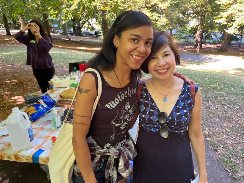 Two writers smiling--one wearing a Motörhead shirt and a flannel, the other in a blue dress. Behind, there's a picnic table and someone taking a photo, and TREES.