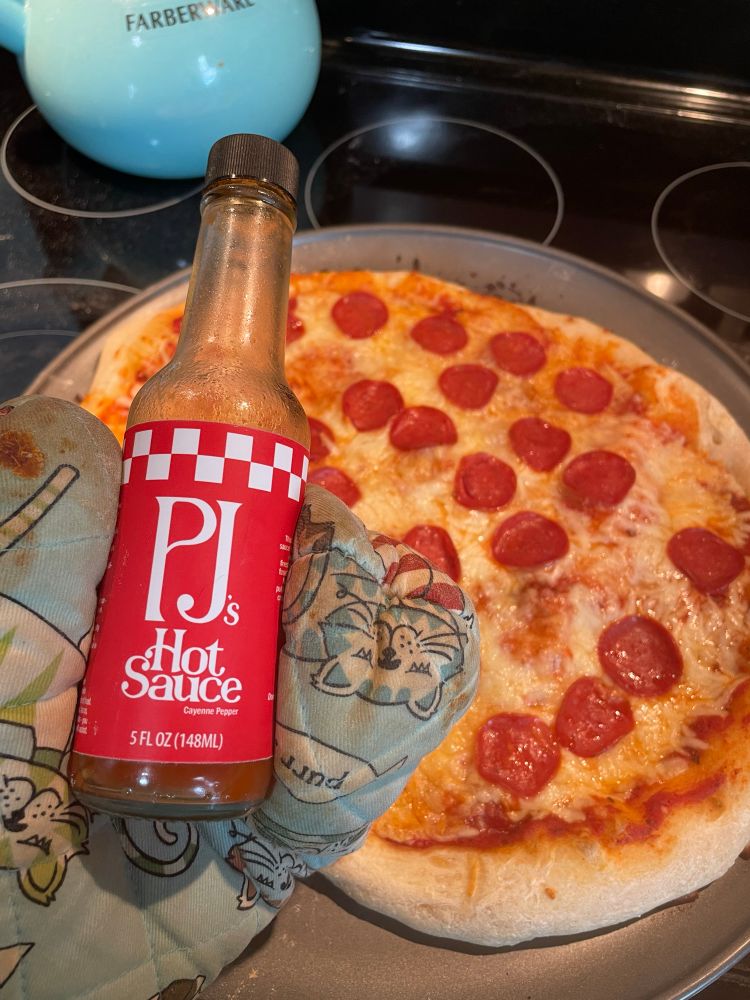 A be-oven-mitten hand holds a nearly empty bottle of Pizza John’s Hot Sauce in front of a homemade pizza. The oven mitt has cats on it as all good oven mitts should, obviously.
