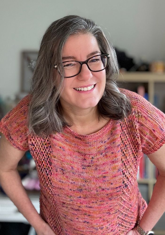 Woman wearing a hand knit short sleeve top with columns of eyelet lace down the right and left sides. The color is pink with multi colored speckles.