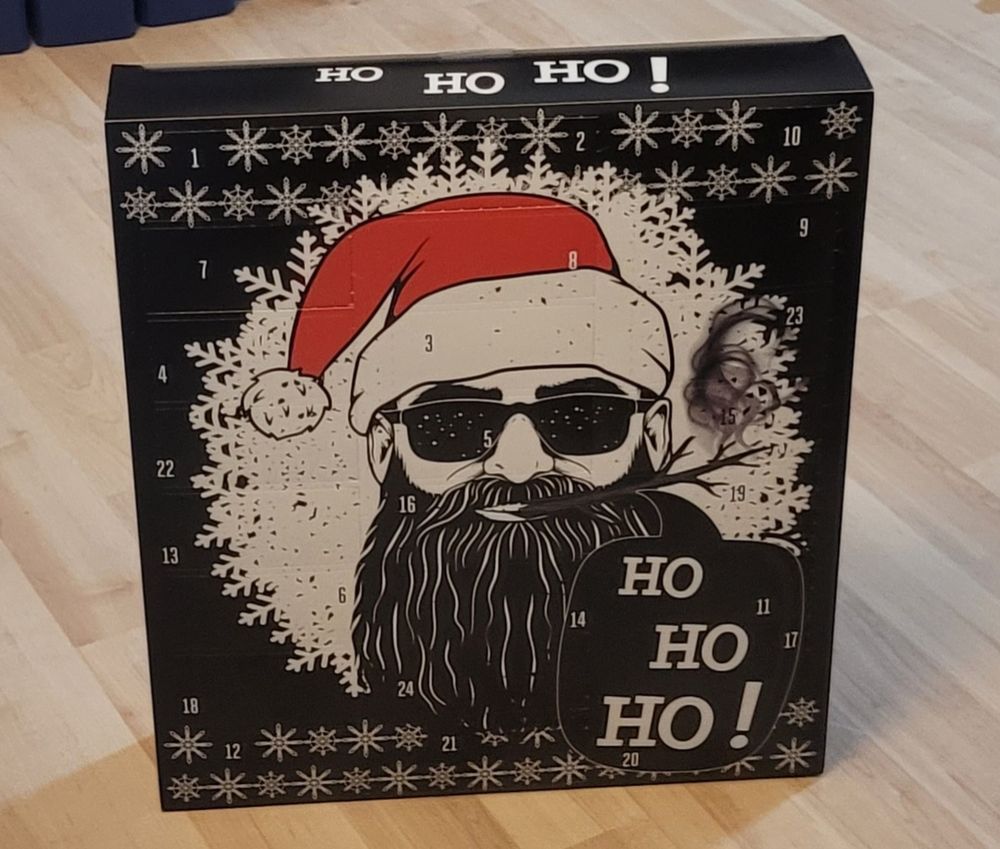 Ein schwarzer Adventskalender. Darauf abgebildet ein Männergesicht mit langem schwarzem Bart, Sonnenbrille und rot-weißer Nikolausmütze.
Daneben ein Schriftzug "Ho Ho Ho"