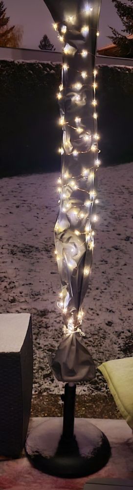Auf der Terrasse steht ein eingepackter Sonnenschirm, Um dessen Hülle ist eine Lichterkette gewickelt. Es ist dunkel und die Lichter leuchten. Im Hintergrund verschneiter Rasen und eine Hecke.