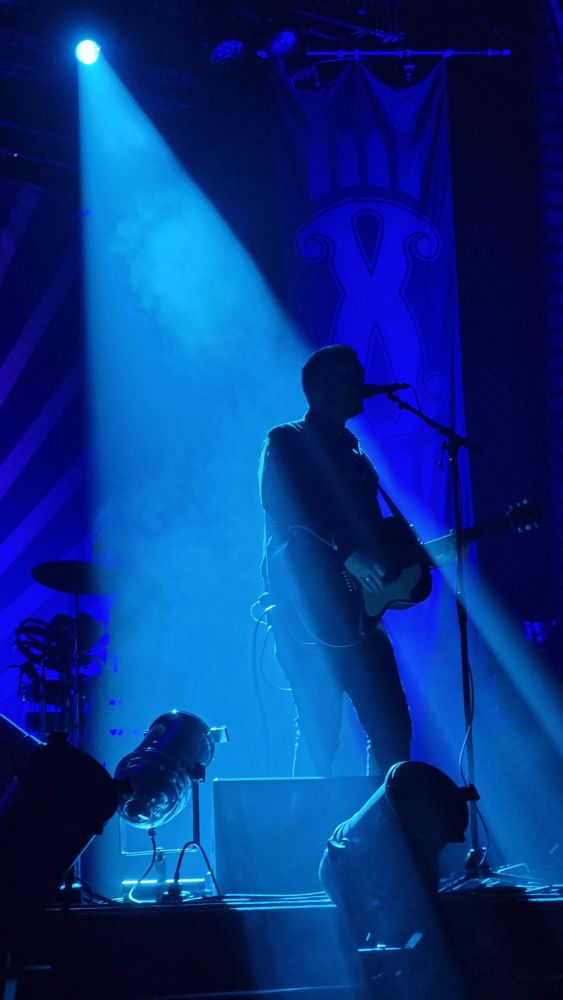 Concert photo: full body shot of the vocalist, stage in dark blue, he is lighted by a single spotlight from behind, creating a beam in the fog, which is split by the singer's body
