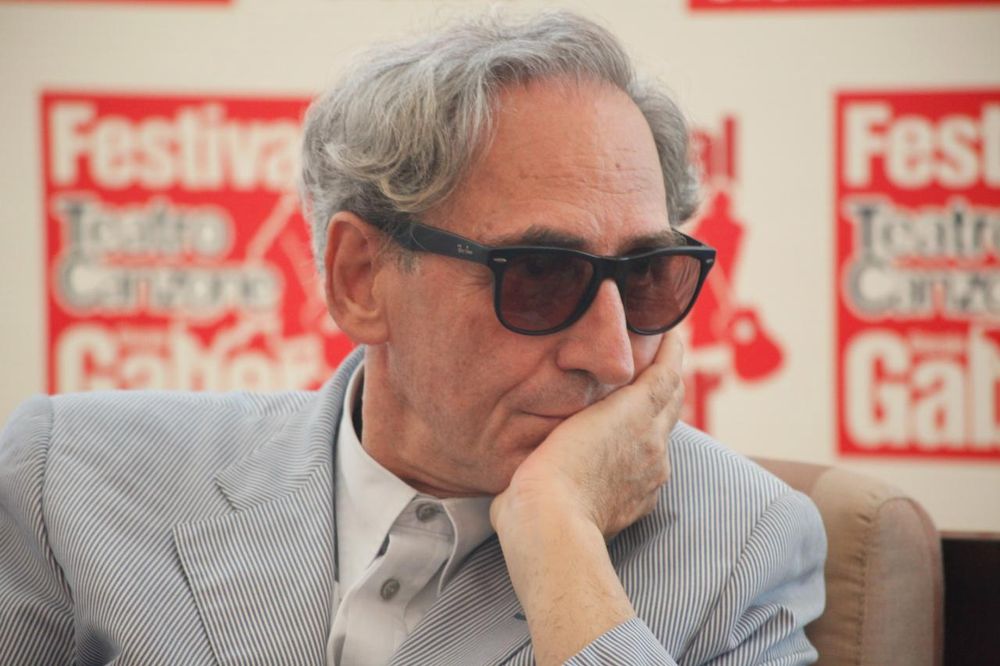 Franco Battiato, pelo canoso ligeramente ondulado, gafas de sol y elegante traje color gris perla, sentado con una mano apoyada en la mandíbula, tapándole parcialmente la boca, en la rueda de prensa del Festival Gaber de 2010. rabendeviaregia • CC BY-SA 2.0
