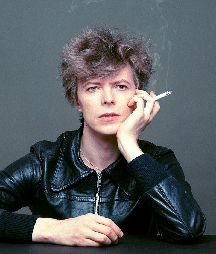 Fotografía de la sesión de fotos de Masayoshi Sukita de donde sale la famosa portada de Heroes. En lo que parece un momento de descanso, Bowie, chaqueta de cuero negro cerrada, una mano apoyada en una mesa, el codo del otro brazo también mientras esa mano se apoya en su mejilla y sujeta un cigarro, mira hacia un lado con gesto indolente y el flequillo levemente despeinado. Masayoshi Sukita.