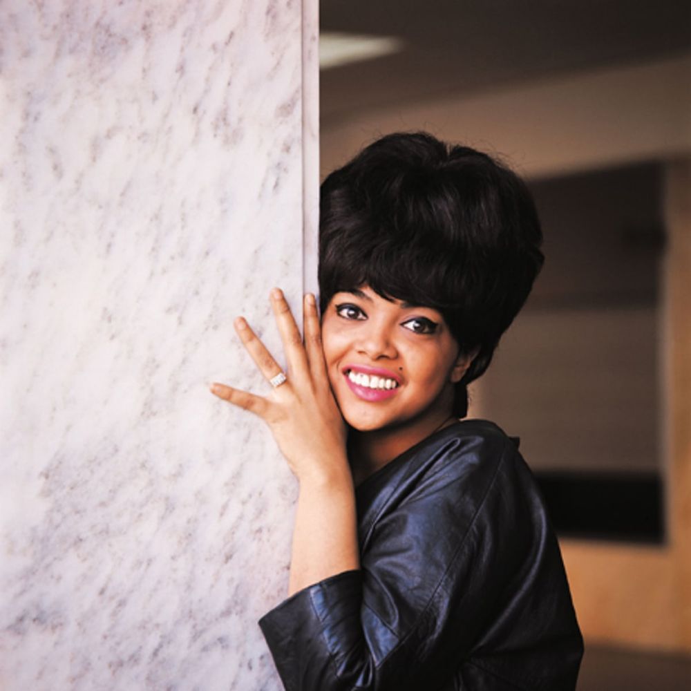 Tammi Terrell, en una fotografía promocional de Tamla Motown. Es una mujer joven, de bonita sonrisa sobre la que vemos un pequeño lunar con un peinado de casco clásico de los 60. Lleva un vestido azul oscuro con brillo y se esconde parcialmente tras una columna blanca, de la que asoma parte de su espalda, su cabeza y una mano con un gran anillo blanco. Discogs.