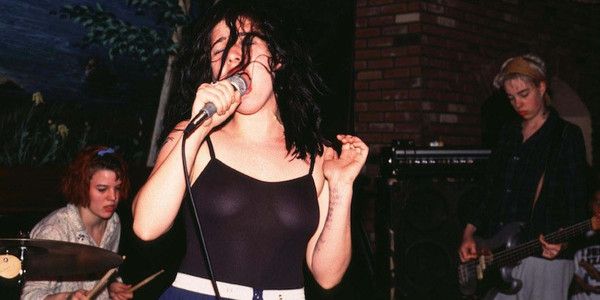 Bikini Kill en concierto a inicios de los 90. De izquierda a derecha, Tobi Vail a la batería, Kathleen Hanna en primer término, con un top negro semi transparente y cantando con el pelo desmelenado y a la derecha, seria y concentrada, Kathi Wilcox, con el pelo corto rubio y tocando el bajo. Discogs.