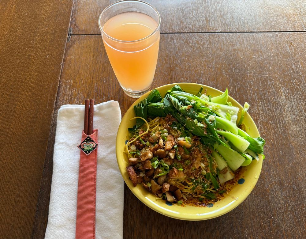 Green bok choy, brown lion’s head mushrooms, yellow noodles, in a bright yellow bowl. Glass of pink grapefruit juice and wooden chopsticks on a white napkin to the side