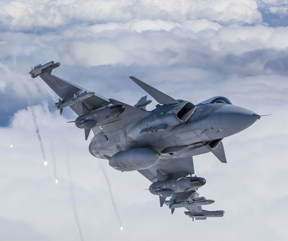 Bilden föreställer det svenska stridsplanet JAS 39 Gripen som flyger i luften bland molnen. Visar mer av undersidan och har släppt flares.