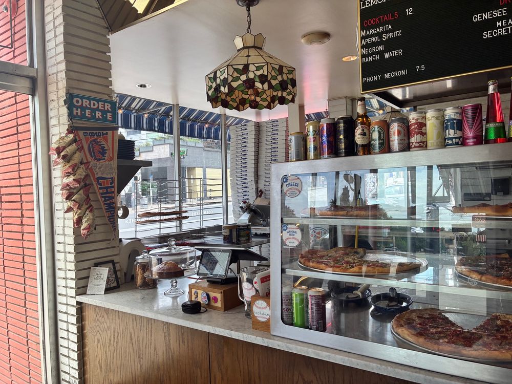 The counter to place your order. Hanging above is a Tiffany-style lamp while a display case of the pizzas to have a slice of are available. On top of the case is a selection of the drinks of choice of beers, sodas, and sparkling water. On the left near the entrance is a 1986 Mets Champions baseball pennant with Mrs. Vick’s kettle chips. The background has large cans of tomatoes, holding racks for pizzas, and towers of pizza boxes. A partially obscured order board listing prices and what is available is in the top right corner