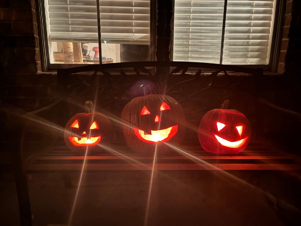 Three pumpkins of varying sizes glowing from the candles within them. The first pumpkin has triangle eyes and nose with buck teeth, the second pumpkin has triangle eyes and nose with a single snaggletooth, and the third pumpkin has triangle eyes with no teeth and no nose