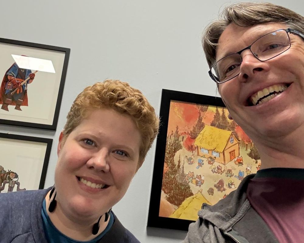 A selfie of a smiling red-haired light-skinned person and a smiling brown-haired light-skinned person with glasses in front of framed artwork.