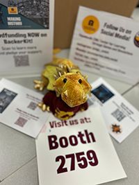 Plush golden dragon in a maroon sweater with a golden 'c' knitted into the front looks up from atop a sign that says BOOTH 2759. It is surrounded by more handouts and signs. 