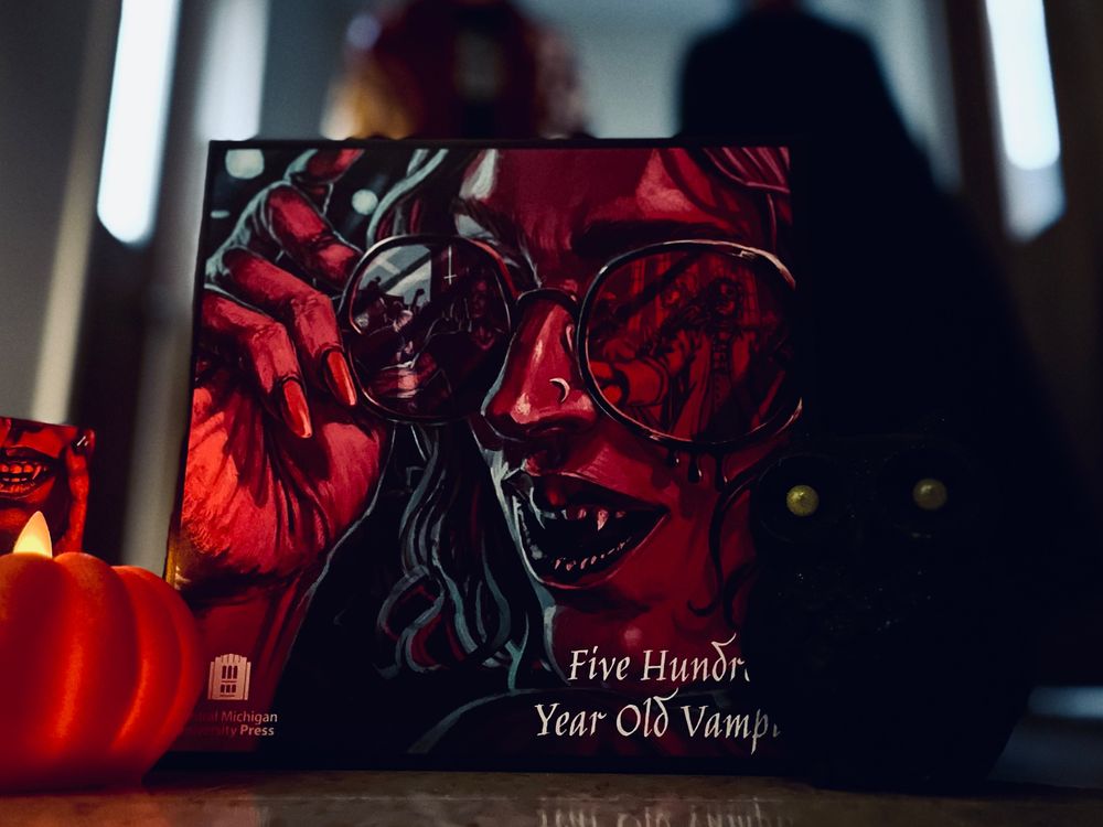 Photograph of Five Hundred Year Old Vampire's box next to a small pumpkin with a flame on top and a black owl. In the background stand two shadowy robed figures.