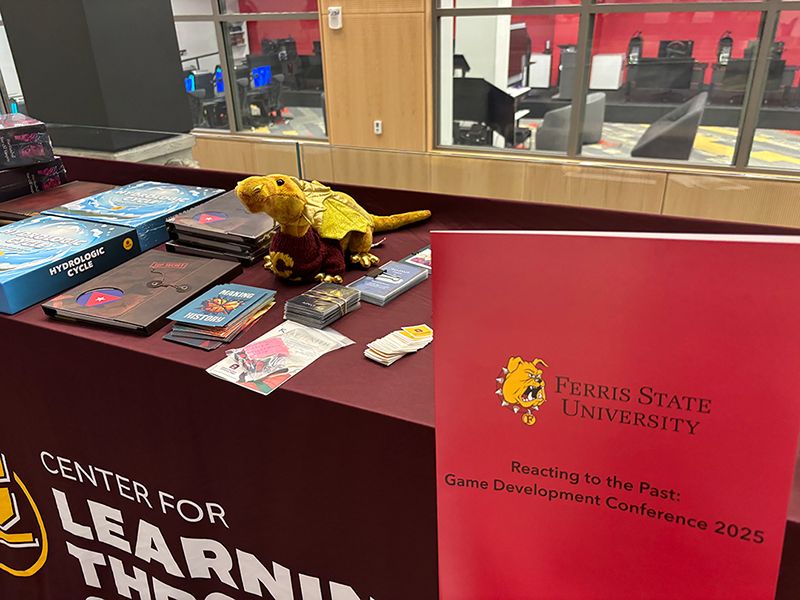 A stuffed golden dragon sits on a table with tabletop game materials. In the foreground is a folder with the words 'FERRIS STATE UNIVERSITY REACTING TO THE PAST: GAME DEVELOPMENT CONFERENCE 2025'.