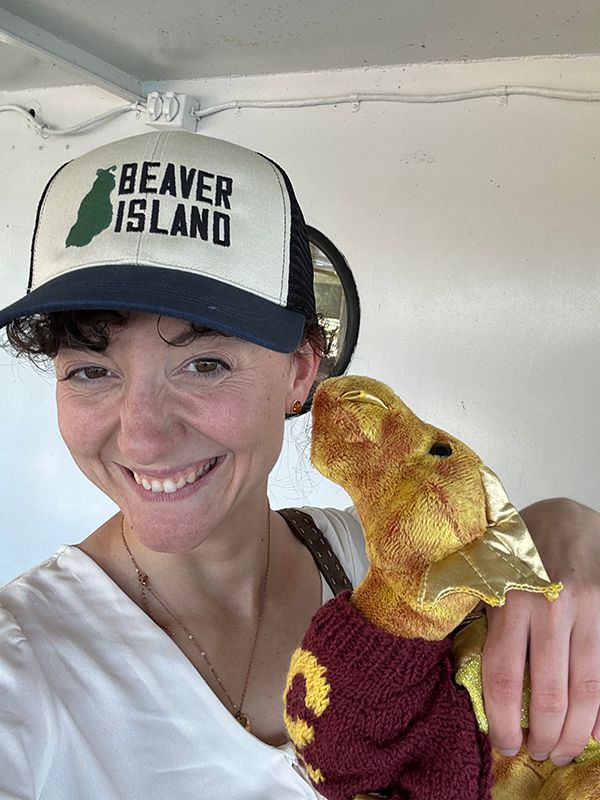 A young white woman with dark hair and a baseball cap which reads BEAVER ISLAND holds the stuffed golden dragon and smiles. They are in front of a white boat surface with a porthole.