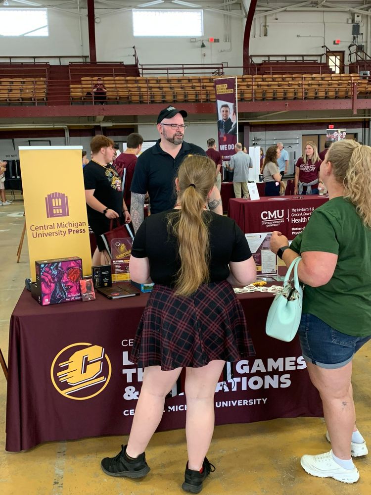 A man stands behind a table marked CENTER FOR LEARNING THROUGH GAMES AND SIMULATIONS with games on the tabletop. He is white with glasses and tall. He is speaking with two young blonde white women.