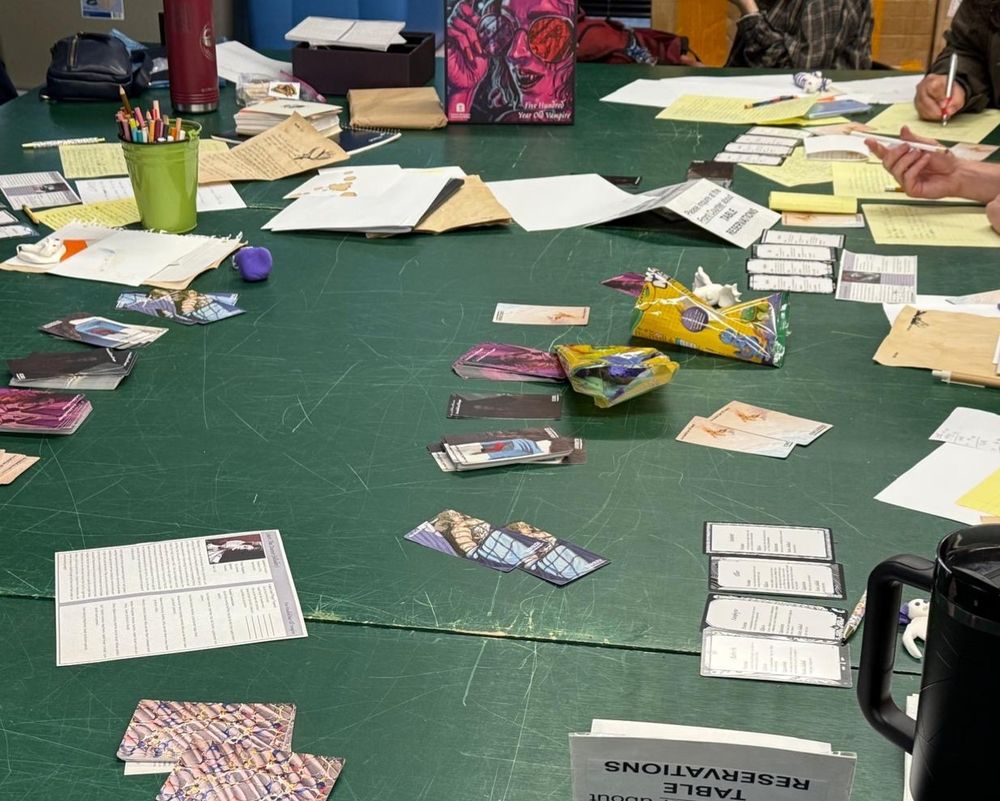 A green tabletop surface covered with papers%2C packets of clay%2C and cards. The box for Five Hundred Year Old Vampire%2C which is magenta with an image of a vampire smiling over sunglasses%2C is visible.