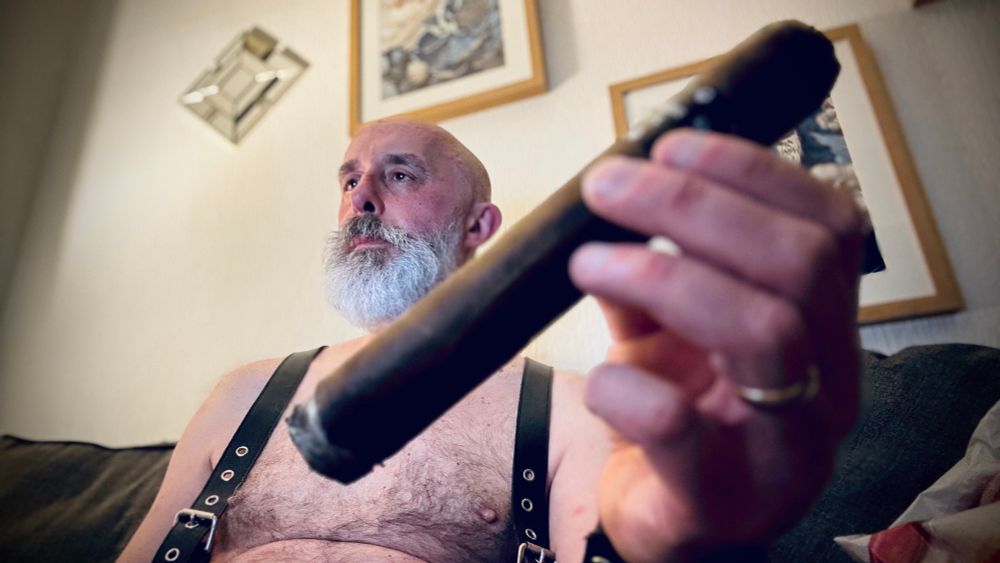Nigel, white bearded, wearing leather suspenders and  is bare chested, and smoking an asylum 8x80 cigar which is in close up 