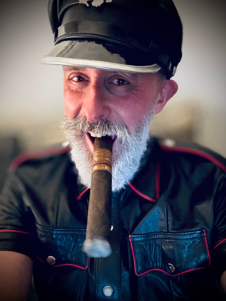 White bearded man, with a large lit cigar clenched in his teeth,wearing a black leather shirt and a Muir cap