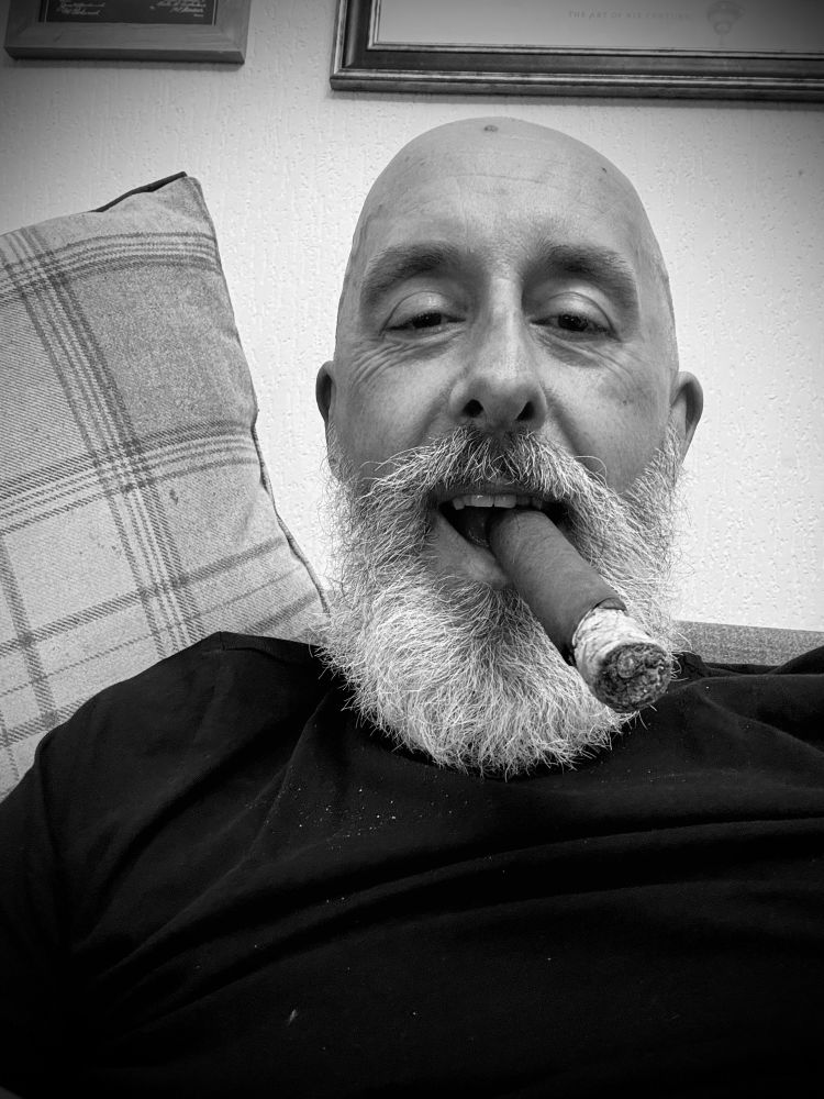 Nigel, white bearded, wearing a black sweater , and smoking large ring cigar seen in close up