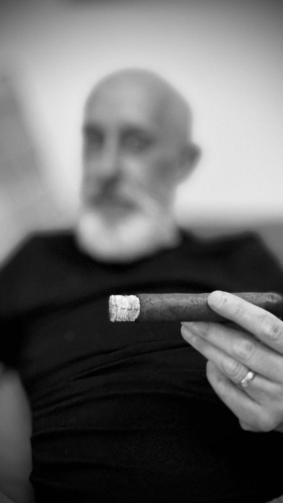 Nigel, white bearded, wearing a black sweater , and smoking large ring cigar seen in close up