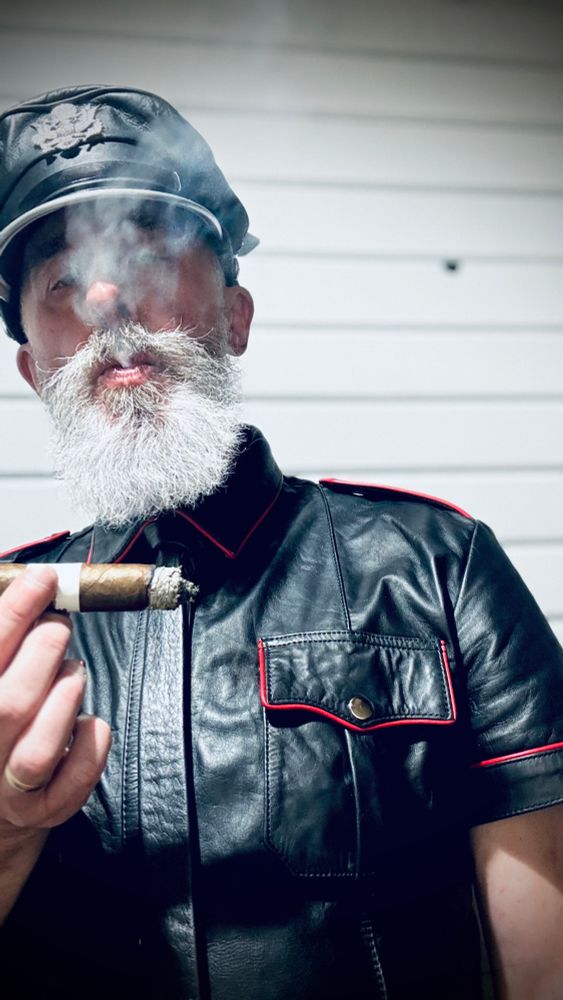 Nigel, white bearded, wearing a black leather shirt with red trim and a Muir cap , and smoking large ring cigar seen in close up